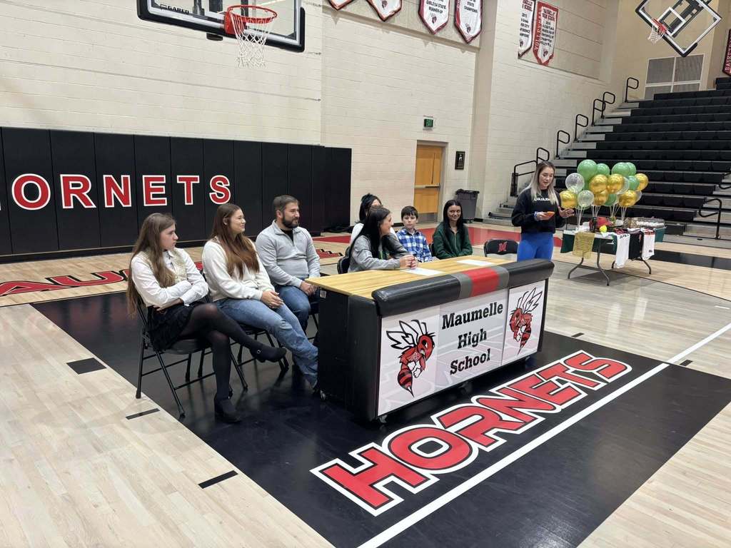 Gracie Keeling in a PJC softball sweatshirt at a table to sign for a college. She is surrounded by others at a gym. The table says Maumelle High School