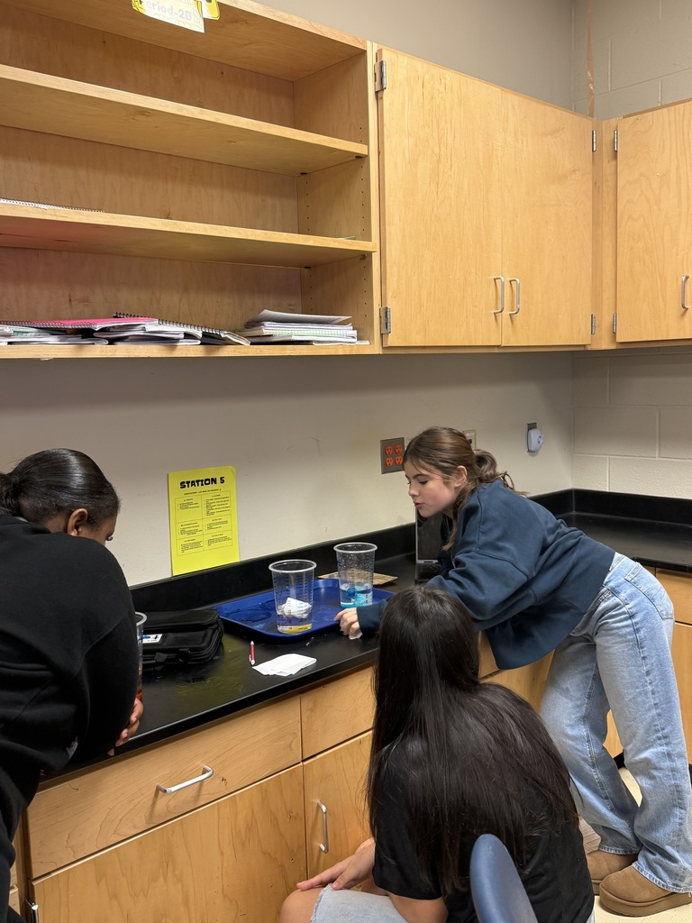 Three students working together in a classroom kitchen with shelves, cabinets, and a safety sign visible.