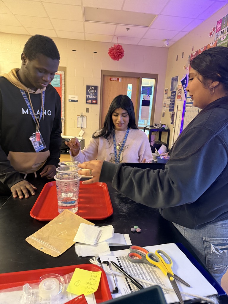 Three people working at a desk in what appears to be an office or school setting. The desk has various supplies and documents on it.