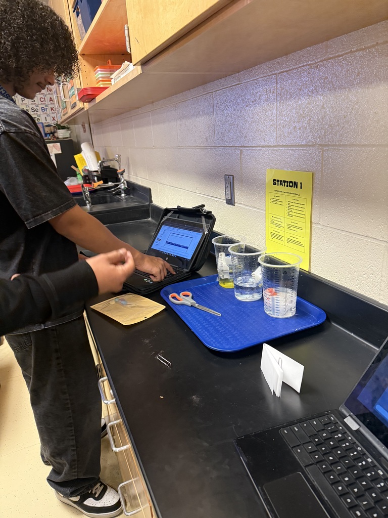 Person working on laptop with various equipment and supplies on counter in what appears to be a laboratory or classroom setting.