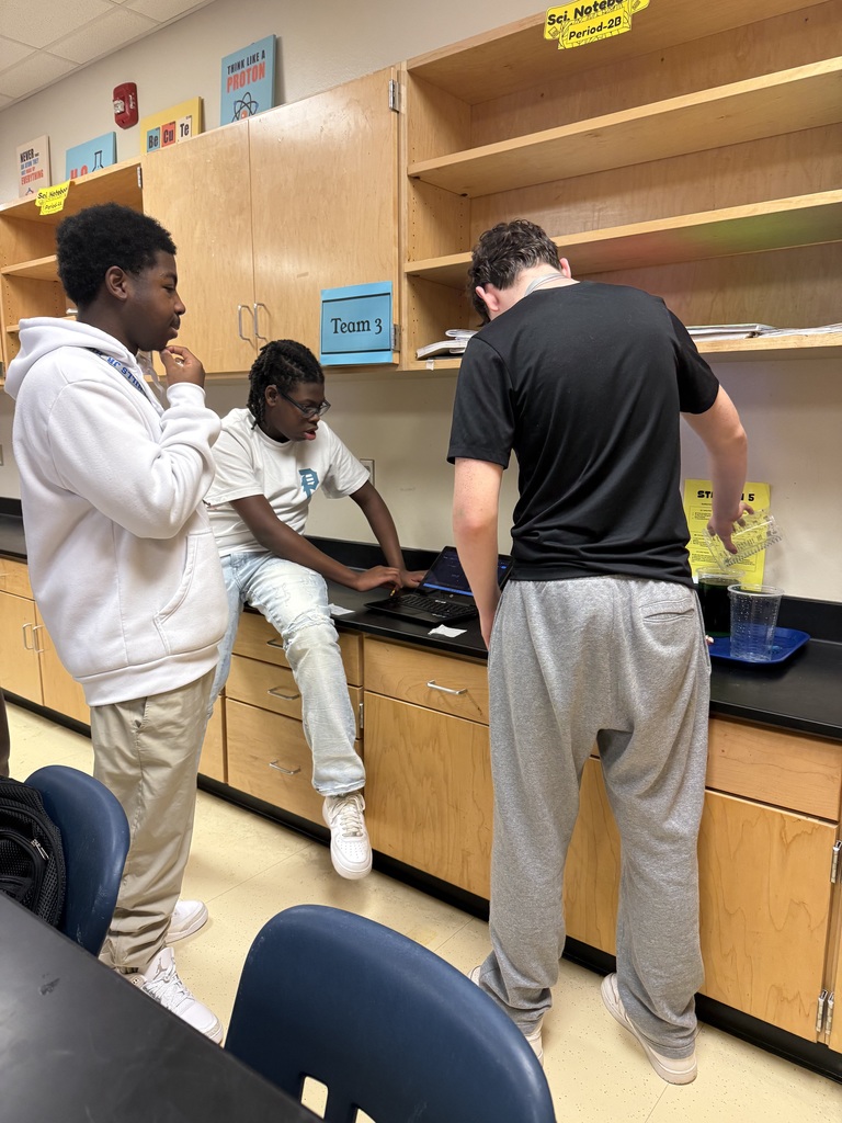 Several students working together at a computer station in a classroom environment with bookshelves and other supplies visible.