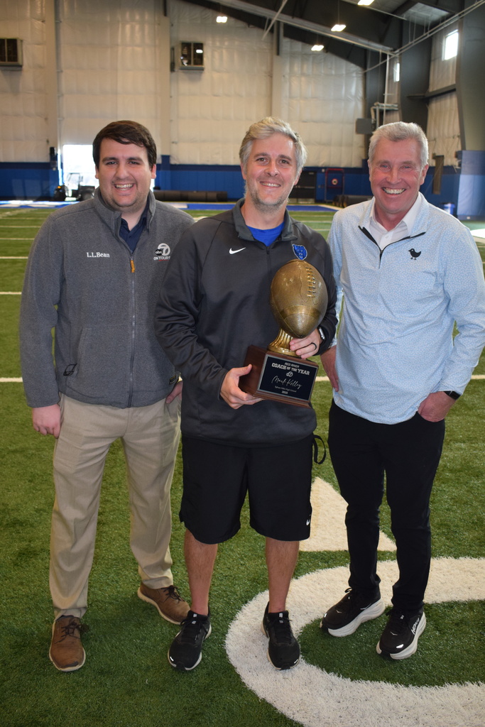 Three men side by side smiling. The middle one is holding a coach of the year trophy.