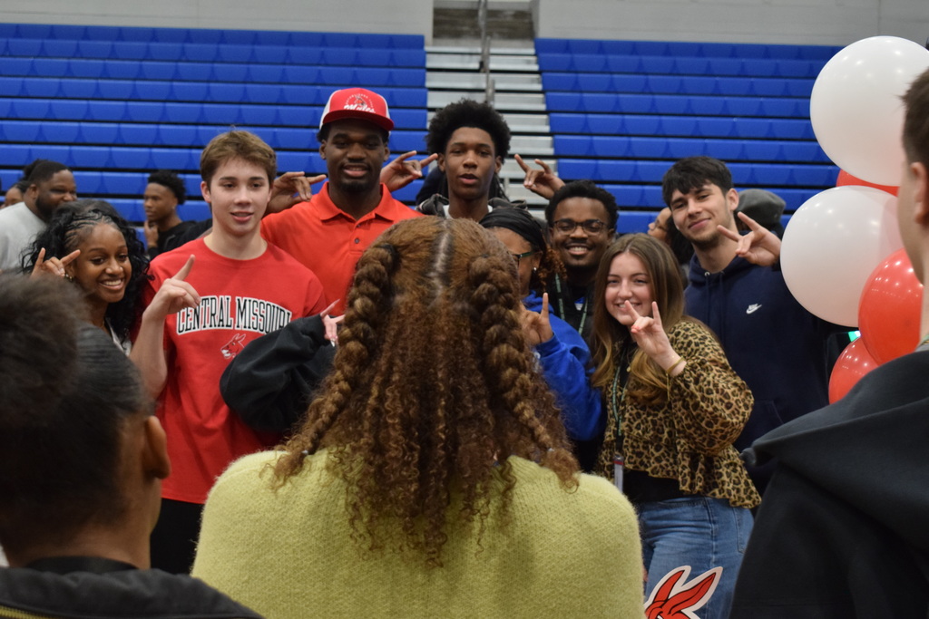 A group of students smile as someone takes a picture of them together. They are making a hand sign where their index finger and pinky are extended while the rest of their fingers are clamped together.