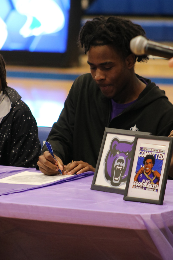 Reggieon Walker signs his letter of intent to play football at UCA. He is sitting at a table with a purple tablecloth and pictures while holding a blue pen.