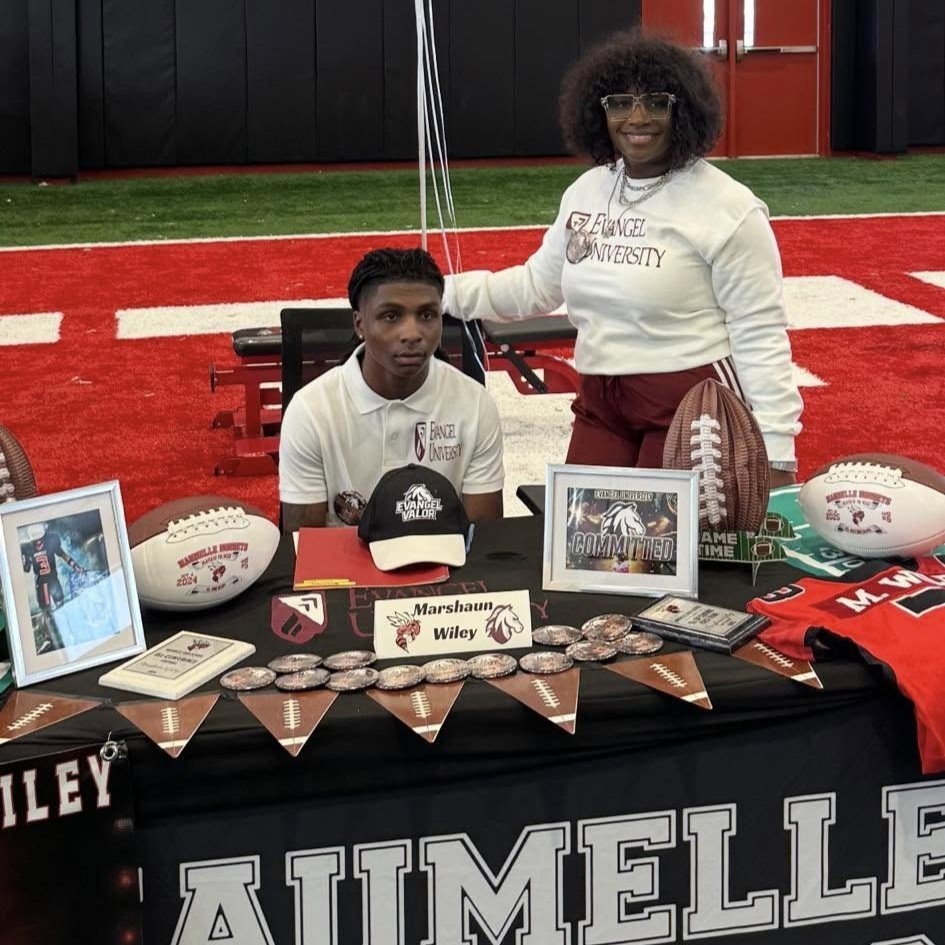 A Maumelle Hornet football player sits at a table with his family smiling. The table says "Maumelle High School". He is about to sign to play at a college