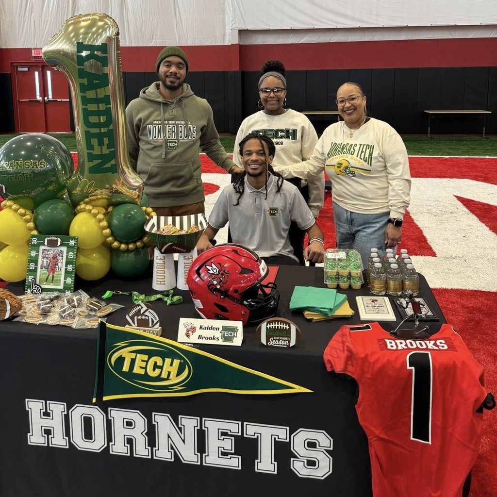 A Maumelle Hornet football player sits at a table with his family smiling. The table says "HORNETS". He is about to sign to play at Arkansas Tech University.