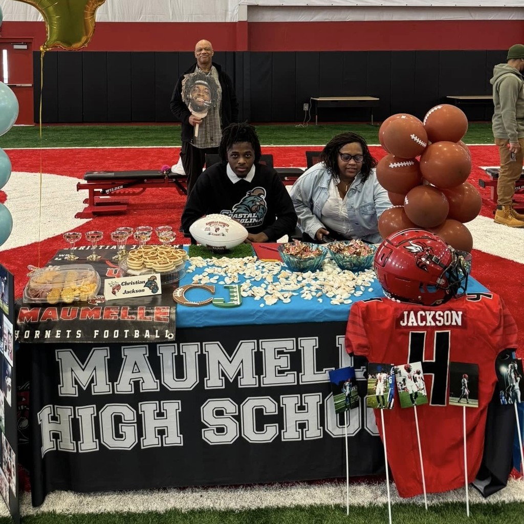 A Maumelle Hornet football player sits at a table with his family smiling. The table says "Maumelle High School". He is about to sign to play at a college