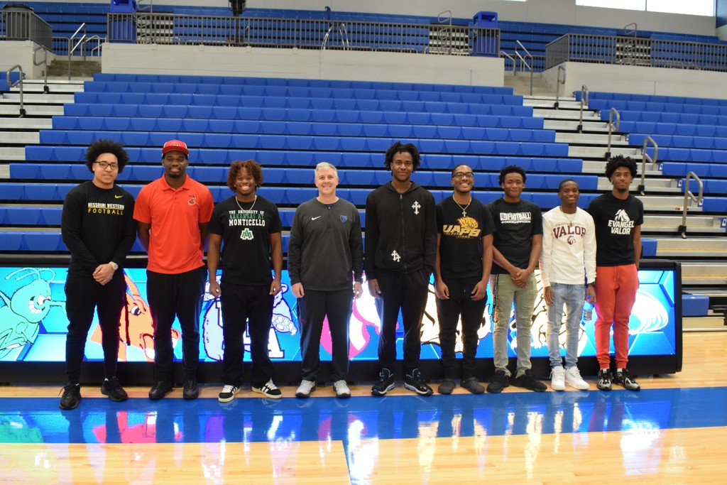 Nine student football athletes and their coach stand together shoulder to shoulder smiling in a basketball arena