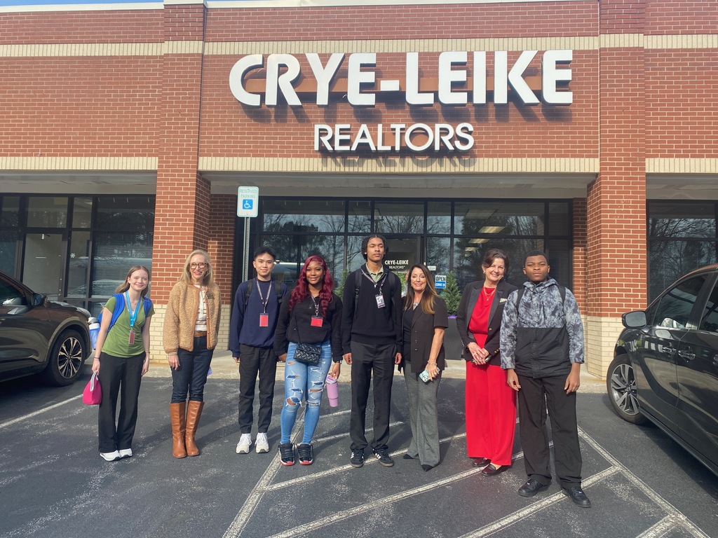 Eight pepole stand in front a strip mall. The business sign behind them says "CRYE-LEIKE REALTORS"