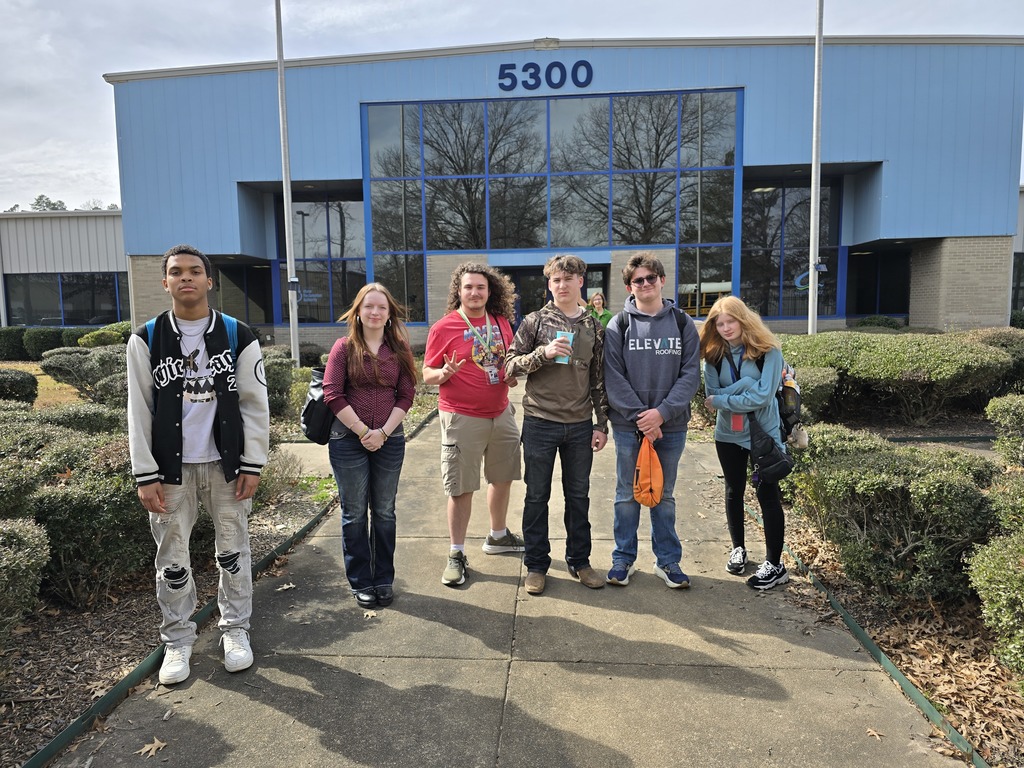 Six people stand in front of a building with shrubbery around them. Most appear to be students