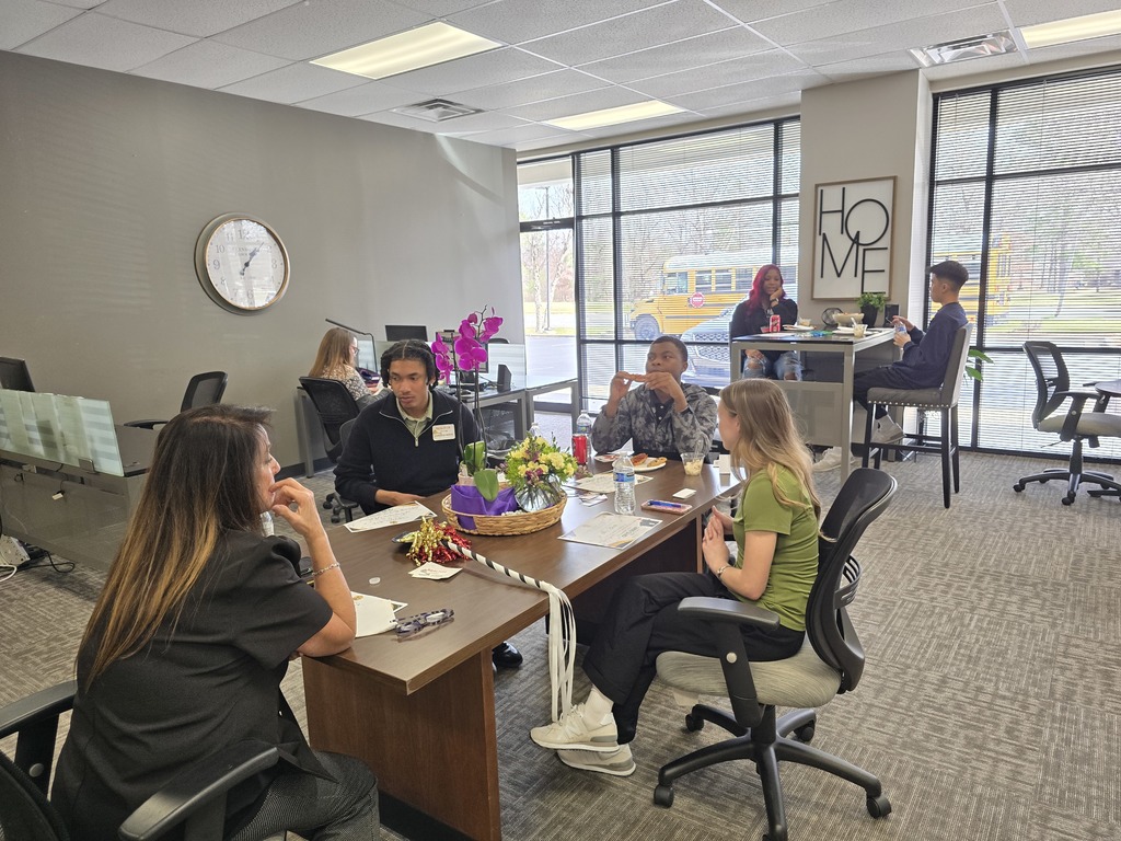 Seven people sit in an office setting. A clock and "HOME" signs are on the walls. A school bus can be seen behind the blinds of the window.