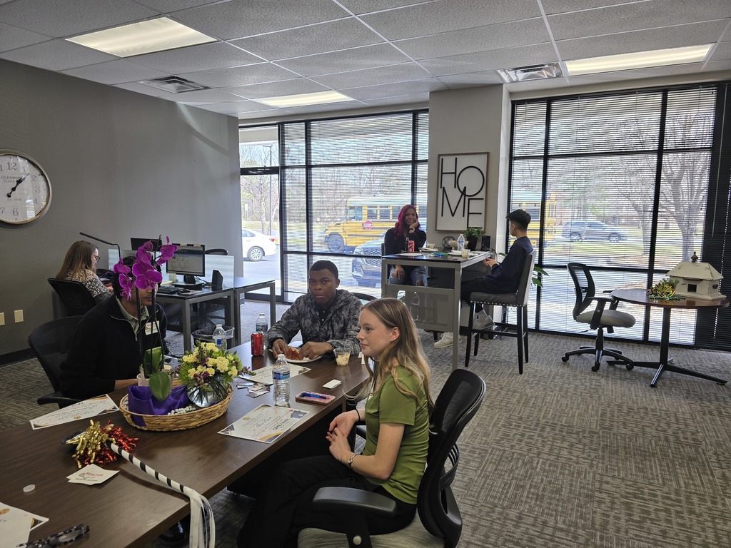 Seven people sit in an office setting. A clock and "HOME" signs are on the walls. A school bus can be seen behind the blinds of the window.