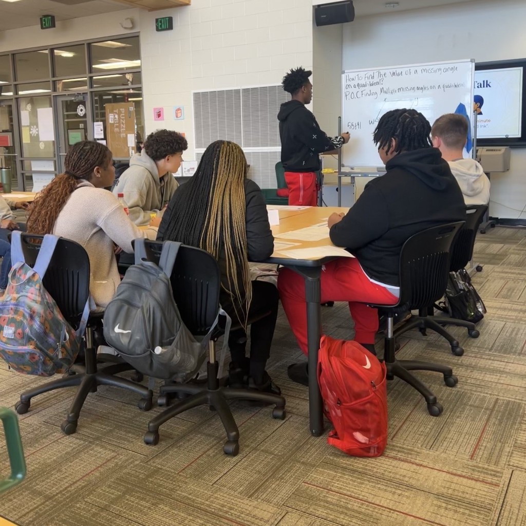 Maumelle High School 9th grade AVID I students showcasing AVID tutorial strategies for all secondary principals and AVID coordinators in the district. One student is using a whiteboard while many look to him in the library.