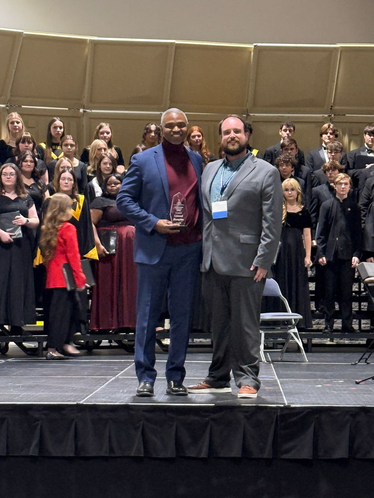 Edmond Hampton holding his choral director of the year award. Another man is beside him and students are in the background.