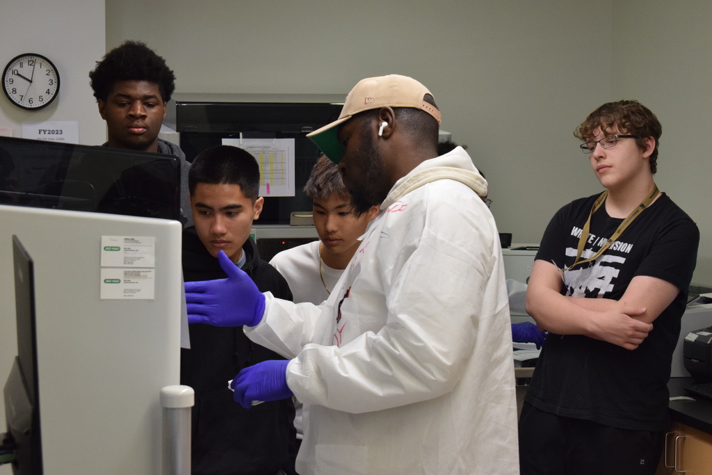 Joe T. Robinson High School Health Science Pathway are learning about lab testing from a a man in a lab coat, ballcap, and ear phones.