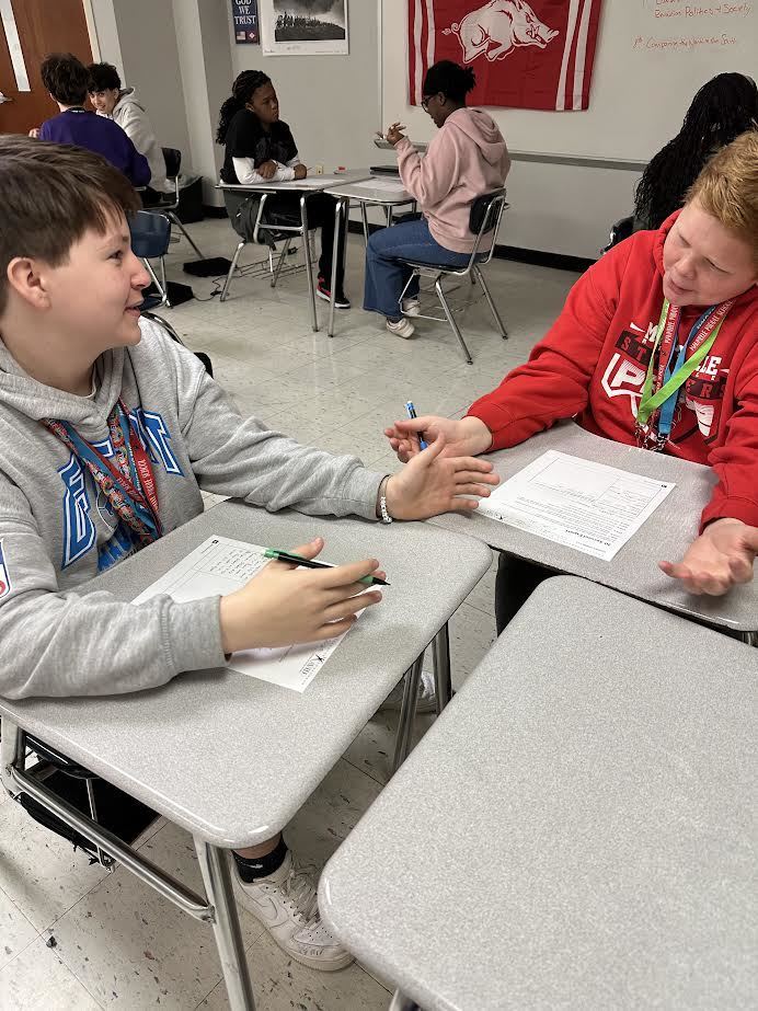 Two students talking with each other about a class project. Others are doing the same in the background of the classroom.