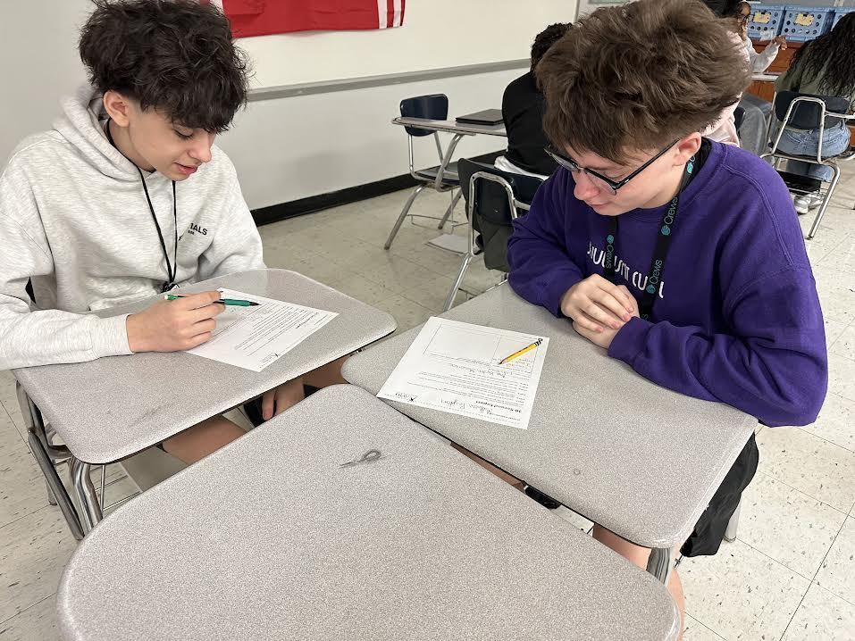 Two students talking with each other about a class project. Others are doing the same in the background of the classroom.