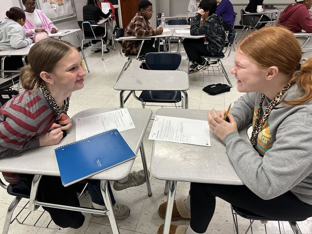 Two students talking with each other about a class project. Others are doing the same in the background of the classroom.