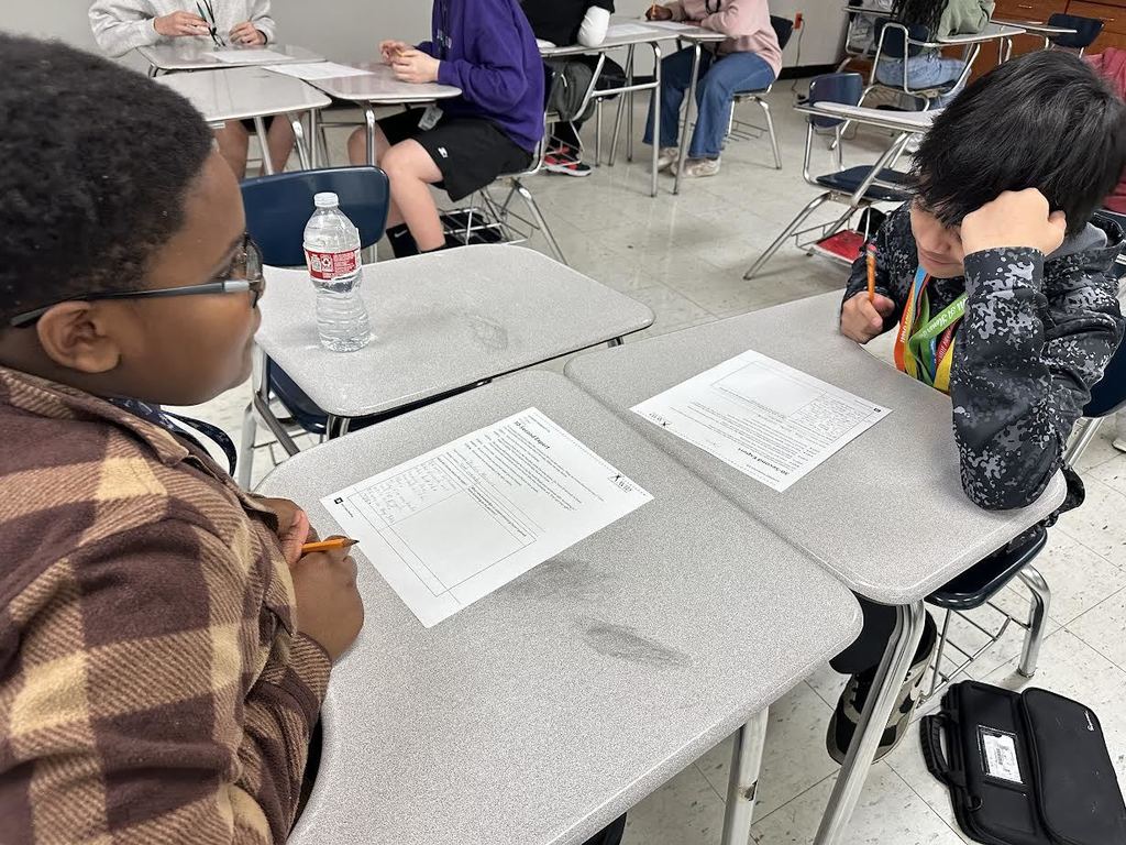 Two students talking with each other about a class project. Others are doing the same in the background of the classroom.