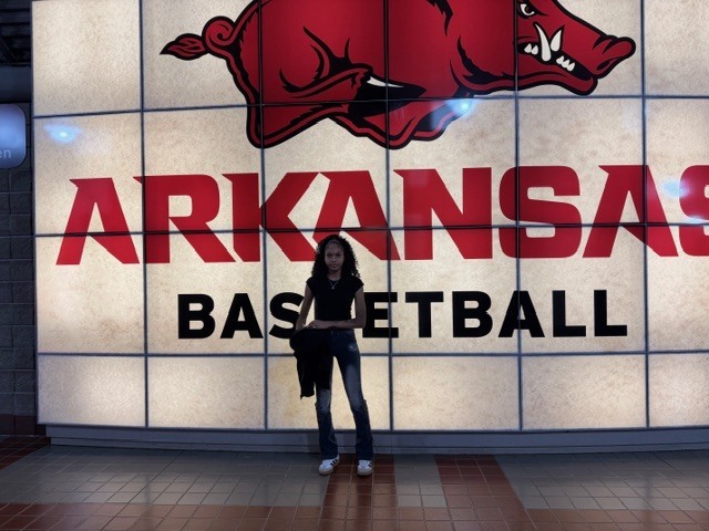 A girl standing in front of a sign that says "Arkansas Basketball" with a Razorback logo above it