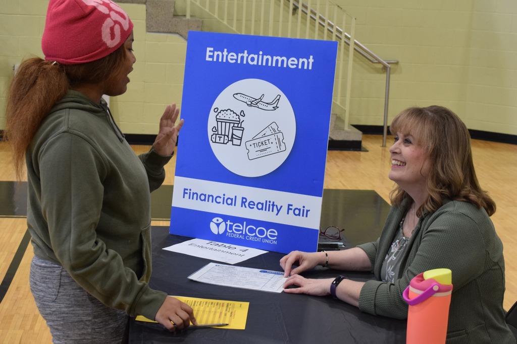 A girl having a discussion with a woman about entertainment while budgeting at a table. Their is a sign and logo for the discussion topic.