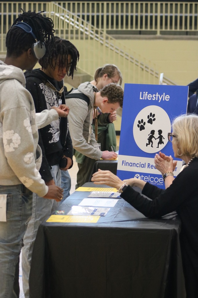 Students having a discussion about lifestyle while budgeting at a table. Their is a sign and logo for the discussion topic.