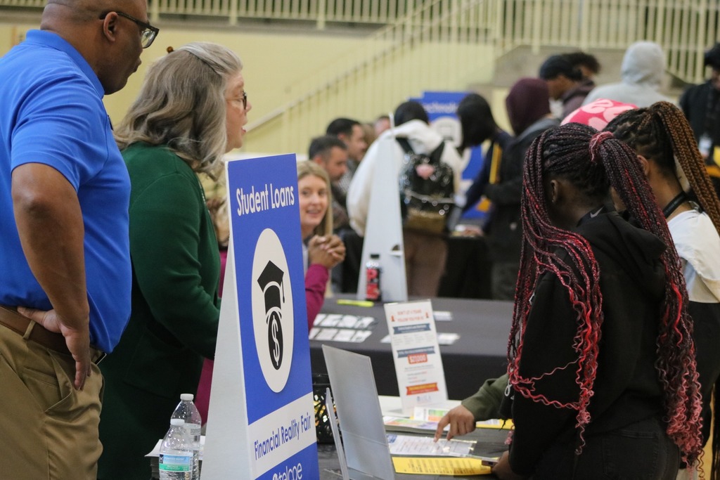 Students having a discussion about student loans while budgeting at a table. Their is a sign and logo for the discussion topic.