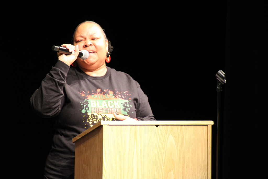 A woman at a podium holding a microphone and saying something. Her shirt says "MILLS BLACK HISTORY"