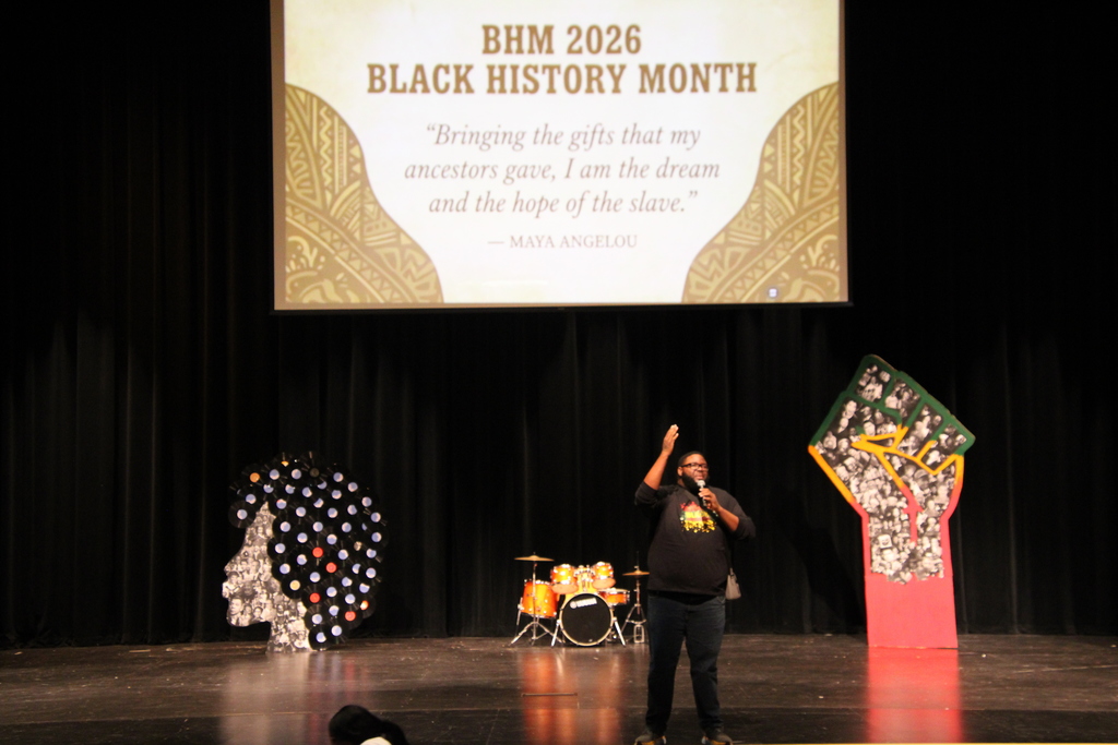 A stage with a teacher and behind him is a projector. The screen says "BHM 2026 BLACK HISTORY MONTH 'Bringing the gifts that my ancestors gave, I am the dream and the hope of the slave.' -Mary Angelou"