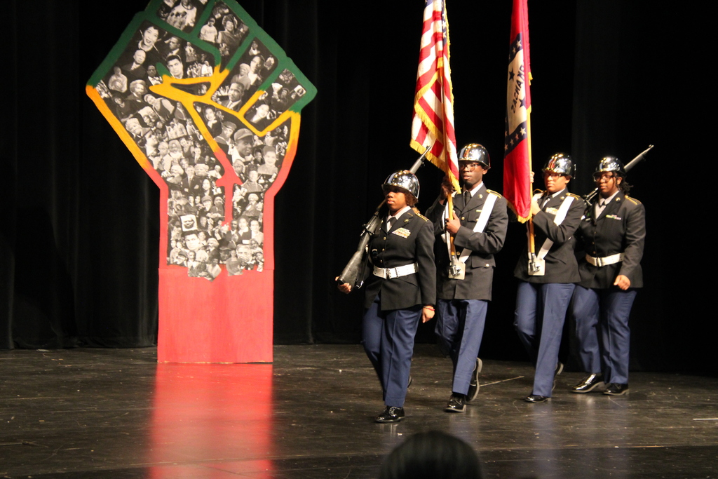 A JROTC group marching with two rifles and two flags by a black power collage