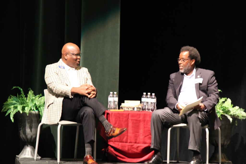 Two grown men having a discussion on a stage. Both are sitting in chairs and have a table with water bottles beside them.