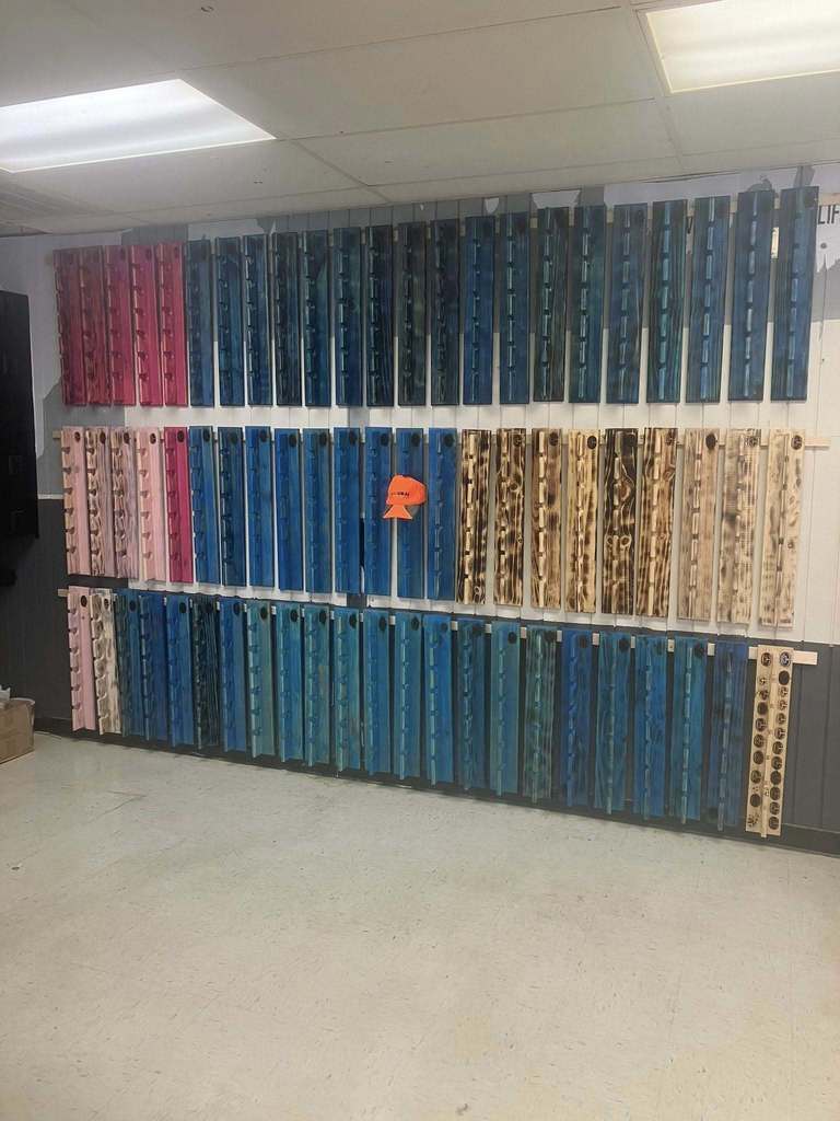 Many hat racks hang on a wall. Some painted blue, others pink, some unpainted. A neon orange hat hangs on the center of the center hat rack