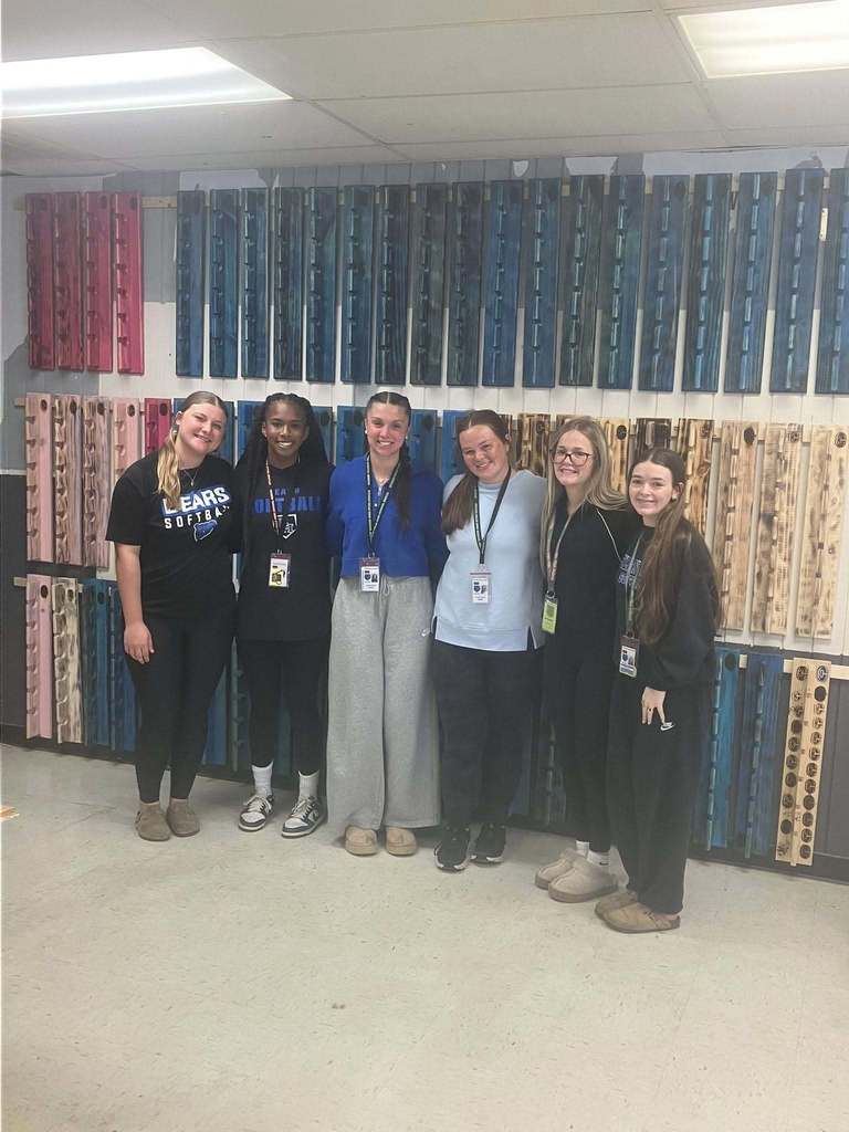 Six girls posed side by side in front of many hat racks.