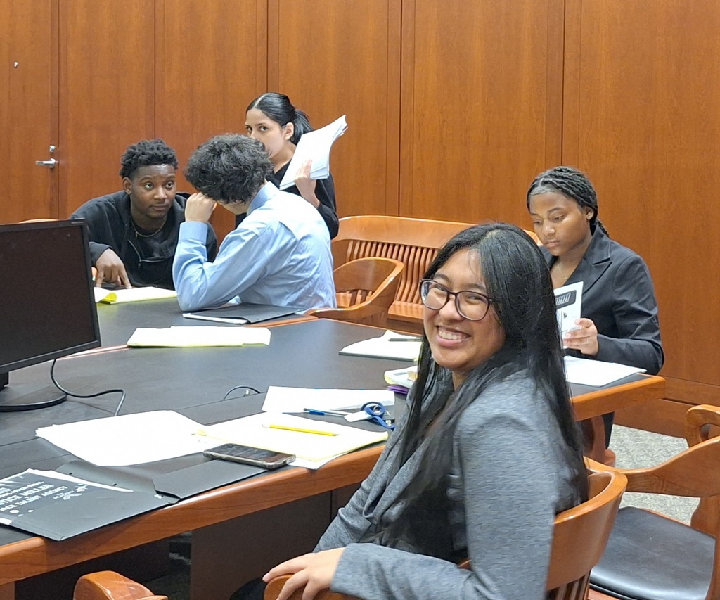 Students in a courtroom. They are sitting at a table discussing and studying. The girl in the foreground is smiling for the camera.