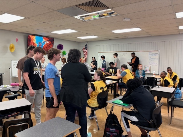 A classroom scene where students and teachers are gathered around the team members in yellow jerseys, chatting and smiling during the sendoff event.