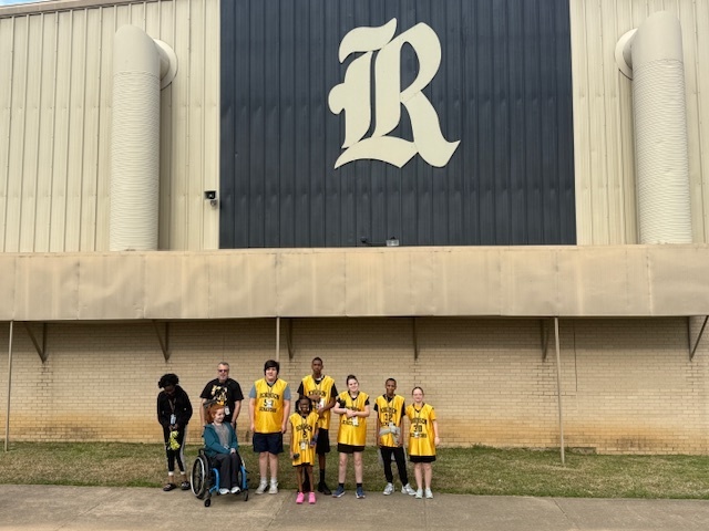 A group of students, mostly wearing yellow Robinson Senators jerseys, and an adult posing for a team photo outside a building under a large "R" logo.