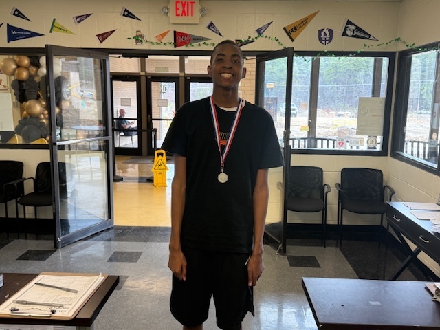 A young man smiling proudly while wearing a silver medal around his neck, standing indoors near a set of glass doors.