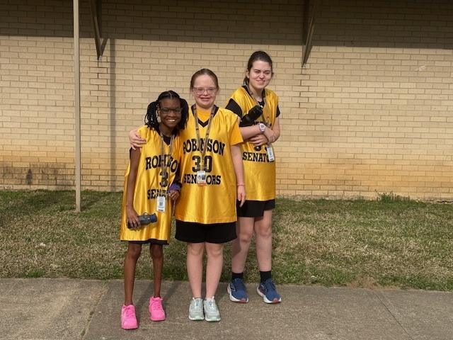 Three smiling students wearing yellow Robinson Senators jerseys posing together outside a brick building.