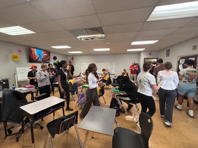 Students in a classroom clapping and giving high-fives to a group of students wearing yellow jerseys during a sendoff celebration.