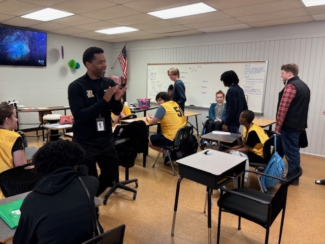 A man wearing a black Robinson pullover claps for the students wearing yellow jerseys who are seated at their desks in the classroom.