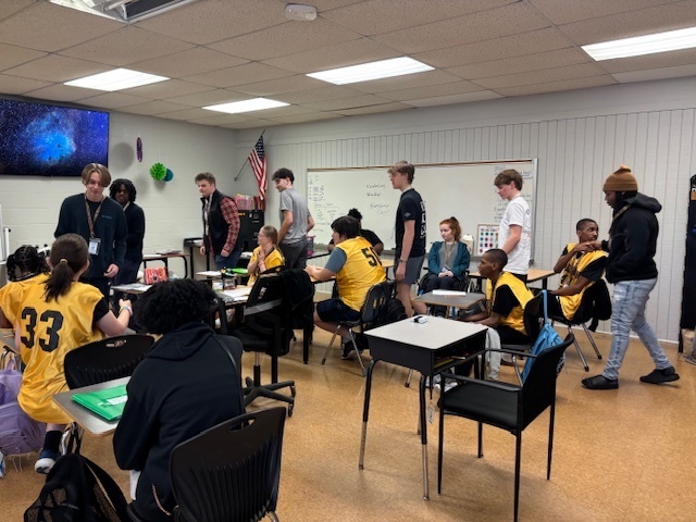 Students wearing yellow jerseys with black numbers sitting at desks in a classroom while their classmates stand around them interacting and celebrating.