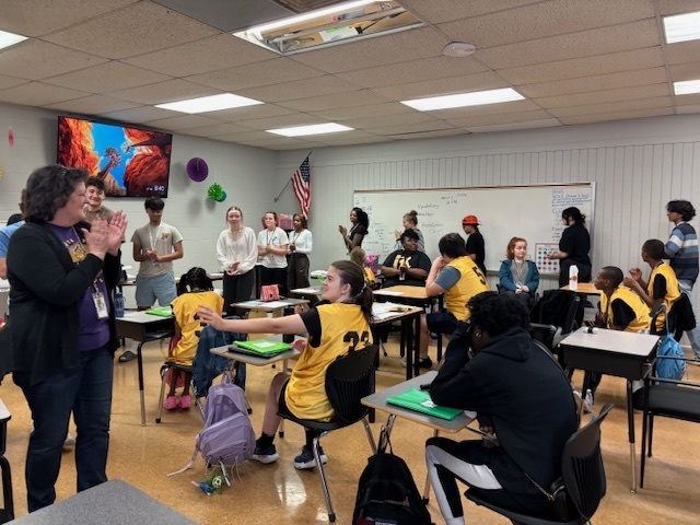 A woman in a purple shirt claps enthusiastically in a classroom filled with students, including several team members wearing yellow jerseys who are smiling.
