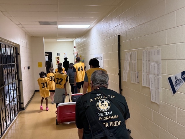 A group of students wearing yellow jerseys with black numbers walking down a school hallway pulling red rolling coolers, followed by an adult wearing a black Robinson Senators shirt.