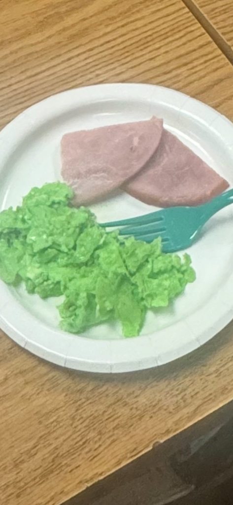 A white paper plate on a wooden desk contains a serving of bright green scrambled eggs and two slices of ham, served with a teal plastic fork.