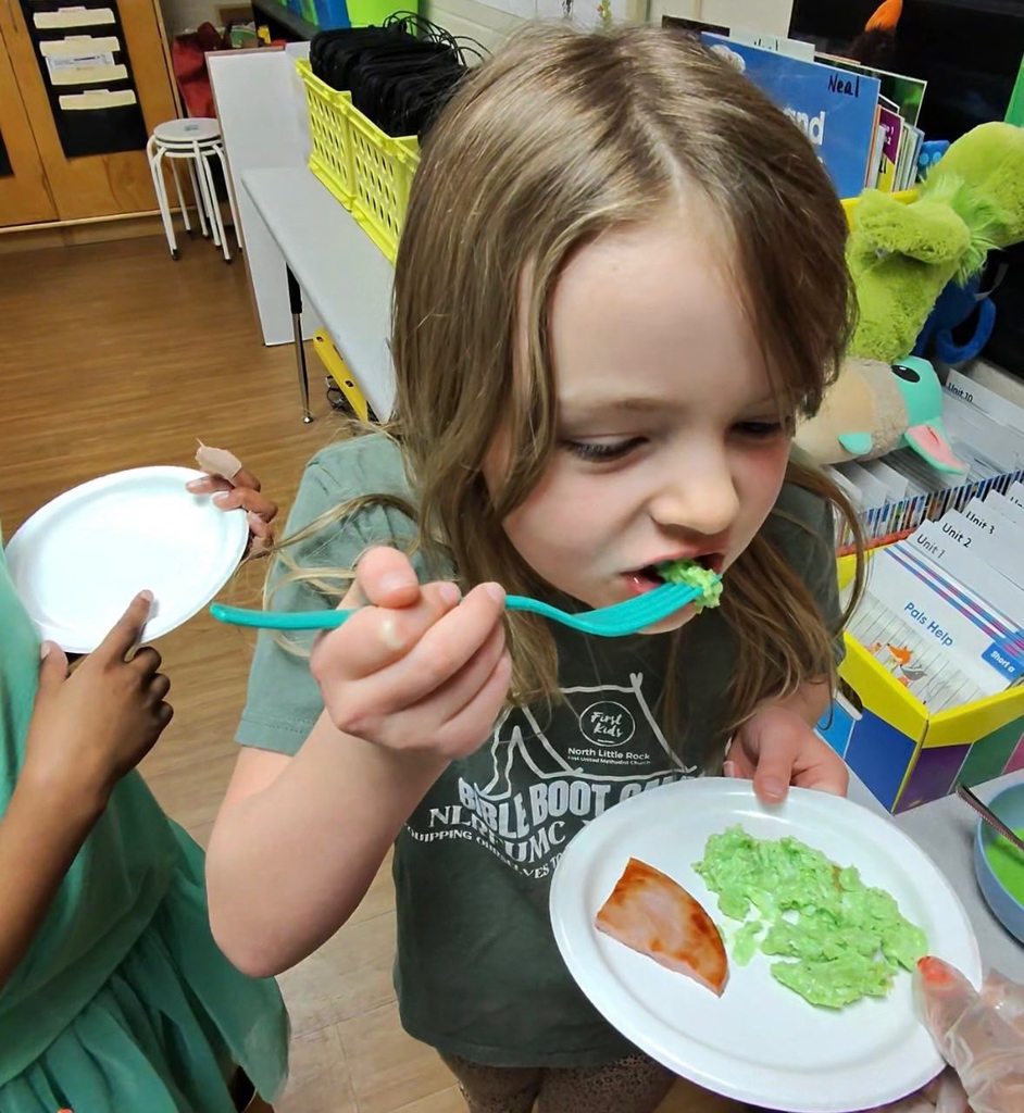 A student in a green t-shirt takes a bite of green scrambled eggs from a white plate that also holds a slice of ham.