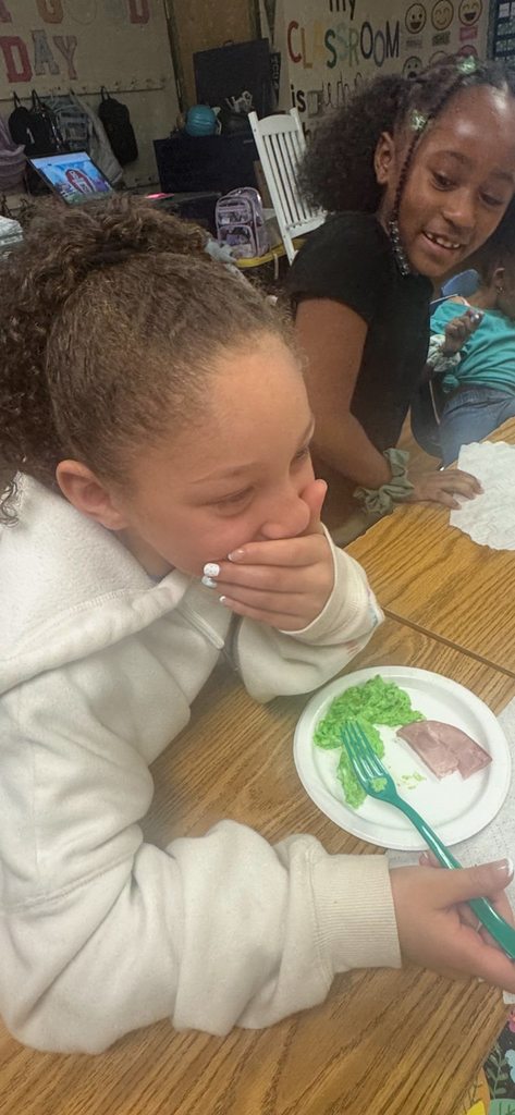 A young student in a white hoodie sits at a desk, looking down at a plate of green eggs and ham with a hand over her mouth, appearing amused or surprised.