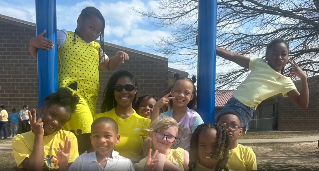 Group photo outside of several students and their teacher outside making a "W" sign with their hands. Some are hanging off playground equipment.
