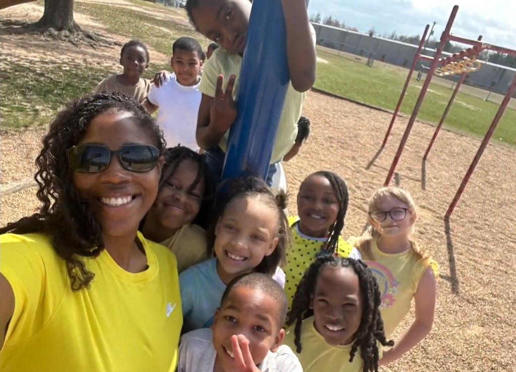 Group photo outside of several students and their teacher outside. One is hanging off playground equipment.