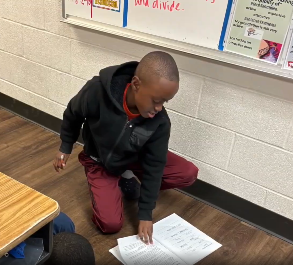 A student kneels on the classroom floor, pointing at a page in an open workbook to read.