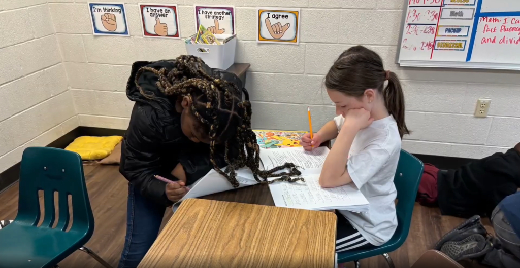 Two students focused on their schoolwork. One girl leans closely over her desk to write, while the other sits next to her, resting her chin on her hand while looking at her assignment.